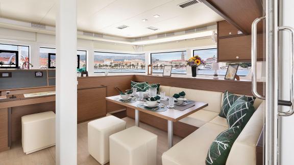 Set dining table and seating area with cushions inside the catamaran, featuring panoramic windows and a coastal town vie