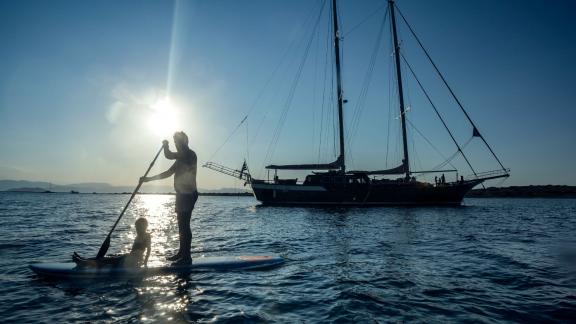 Two people stand-up paddling in front of the gulet Entre Cielos, which is anchored in the background at sunset.