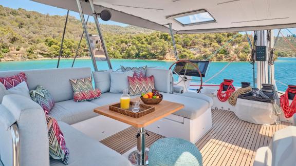 Lounge area with cushions, fruit, and drinks on the catamaran, overlooking the green coast and sea.