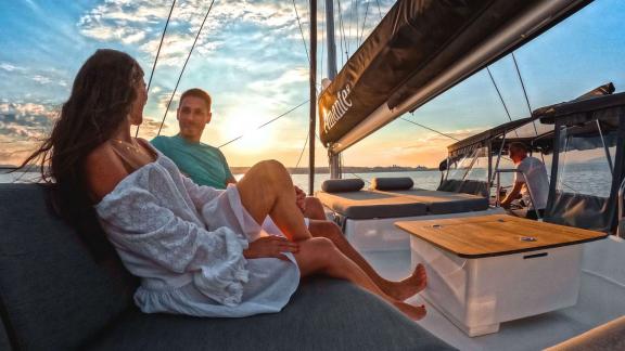 A couple sits relaxed on a catamaran as the sun sets over the sea.