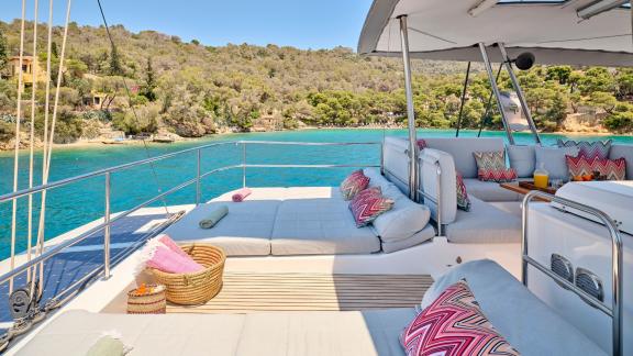 Spacious sun deck of the catamaran with comfortable loungers, cushions, and a view of the green coastline.