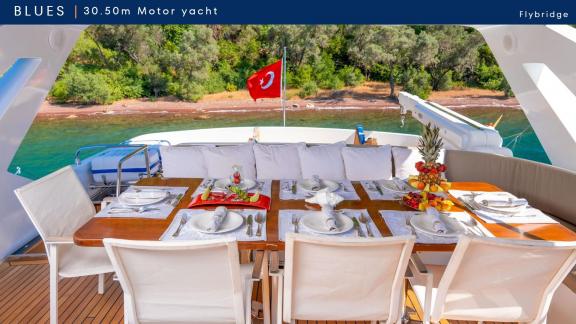 Blues motoryatın flybridge alanındaki masa servisli. Yat kıyıya yakın konumda demirlemiş.