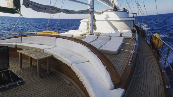 Deck of a large sailing yacht. In the foreground is a comfortable, white upholstered seating area with a wooden table.