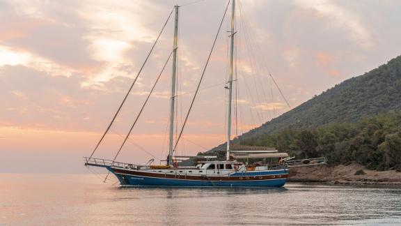 Gulet Gozdem The One at anchor in Bodrum, blue hull, wooden panelling, coastal landscape at sunset.