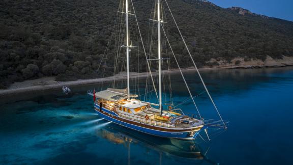 Gulet Gozdem The One beleuchtet vor Anker bei Nacht, blaues Wasser, umgeben von grüner Küstenlandschaft in Bodrum.