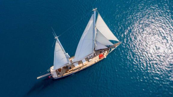 A bird's eye view of a luxurious sailing yacht with white sails sailing on a clear blue sea.