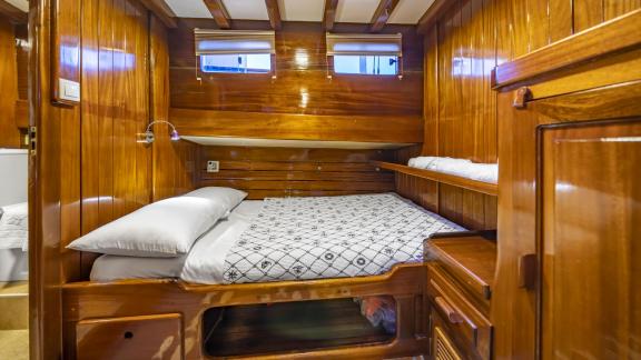 The cabin features a double bed with a patterned cover, surrounded by warm wooden walls and two windows.