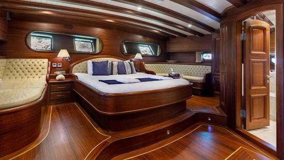 Luxurious cabin with elegant wooden interior, large double bed, seating areas and oval windows for natural light.