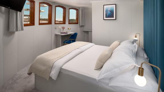 Cozy cabin on the Bellezza yacht with a large bed, desk, and multiple windows offering sea views.