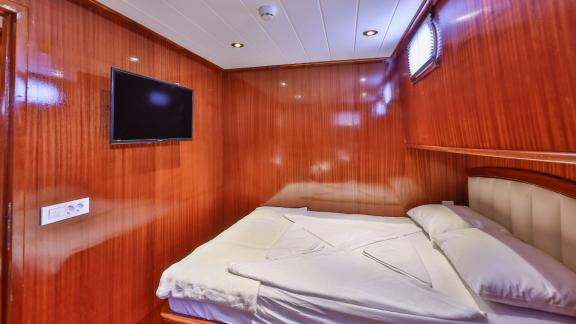 A comfortable cabin on the Gulet Kasapoglu 6 featuring a double bed, wood paneling, and wall-mounted TV.