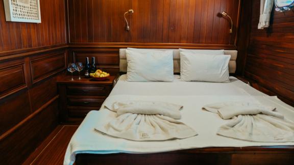 An elegant ship's cabin with a cosy double bed, decorated with towels, a fruit plate and wine on the bedside table.