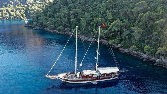 Two-masted yacht anchored in clear blue water off a wooded coast.