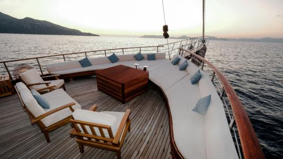 Deck einer Yacht mit einer eleganten Sitzgruppe aus Holz und weißen Polstern. Im Hintergrund ist das ruhige Meer und ein