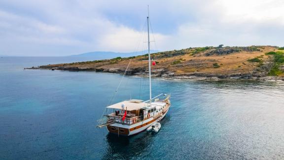 The Gulet Cagan 2 is calmly anchored near a rocky shore, surrounded by clear waters and gentle hills.