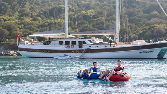 Two people enjoy water sports on floating tyres in front of the gulet Caner 4, which is anchored in a green bay.