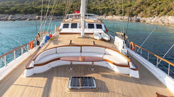 Spacious front deck of the Gulet Caner 4 with seating area and table, surrounded by clear water and green coastal landsc