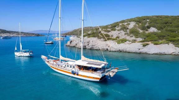 Gulet Cemre Junior is anchored in a bay with blue waters, surrounded by nature.