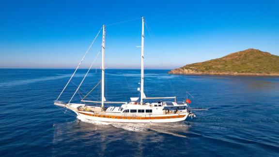 A gulet in Bodrum sails across the calm sea under clear skies, with a green island in the background.