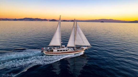 Gulet glides over the calm sea in Bodrum at sunset, with mountain silhouettes in the background.