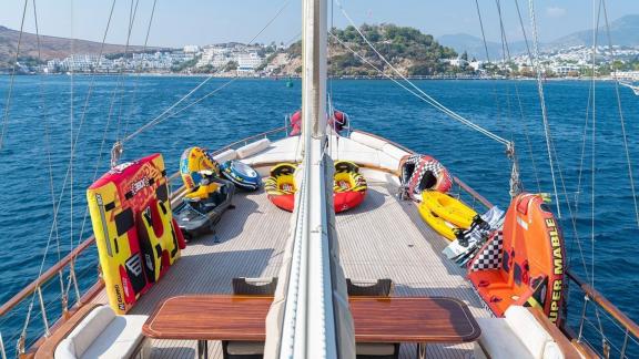 The deck of a gulet is equipped with various water toys and floating tyres, with a coastal town in the background.