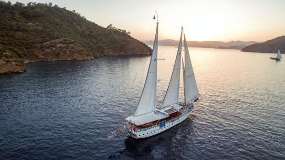 Luxuriöse Gulet Duramaz mit 6 Kabinen segelt bei Sonnenuntergang vor der Küste von Fethiye, umgeben von bewaldeten Hügel