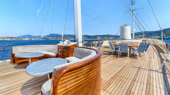 Oberdeck der Elara 1 mit eleganten Holzmöbeln, runden Tischen, Sitzgruppen und Blick auf das Meer und die Hügel.