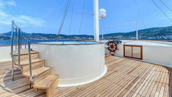 Holzdeck mit Treppe zu erhöhtem Bereich, Steuerrad und weitem Blick auf Meer und Küste.