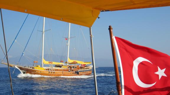 Under sail on the open sea, accompanied by the Turkish flag in the foreground. The ship, with its characteristic yellow