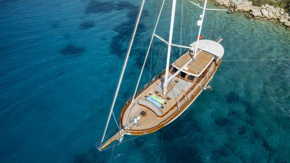 Luxurious wooden sailing yacht with white sails off the coast in clear, turquoise-coloured water.