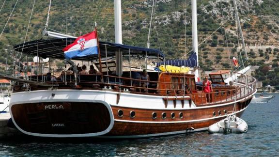 Die Gulet Perla liegt im Hafen von Šibenik vor Anker, mit der kroatischen Flagge gehisst, umgeben von grüner Küstenlands