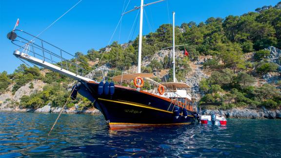 The Gulet Rüya anchored by a wooded coastline with clear blue waters and two dinghies.