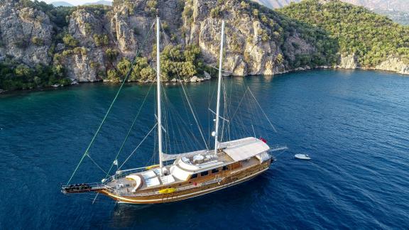 Eine große, elegante Segelyacht, die in einer ruhigen, türkisfarbenen Bucht vor Anker liegt. Im Hintergrund sind steile,