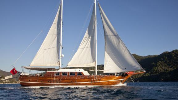 Große Segelyacht mit vollen weißen Segeln, die auf offenem Wasser fährt. Die Yacht hat ein elegantes Holzdesign und träg