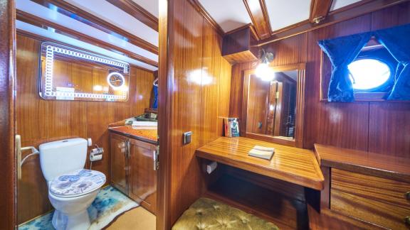 Cabin on a sailing yacht with wooden panelling, bathroom with toilet and washbasin, desk with mirror and small window.