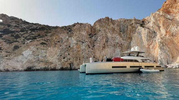 The Lagoon Catamaran is calmly anchored in clear blue waters in front of an impressive cliff, available for charter in G