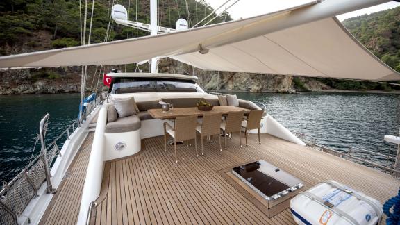 The elegant deck of the Gulet Kayhan 3 with dining area and cosy seating in front of an idyllic coastline.