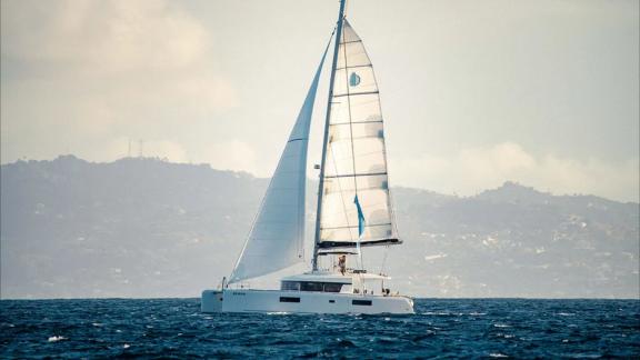 Akana sails under full canvas on open sea with hilly coastline in the background.