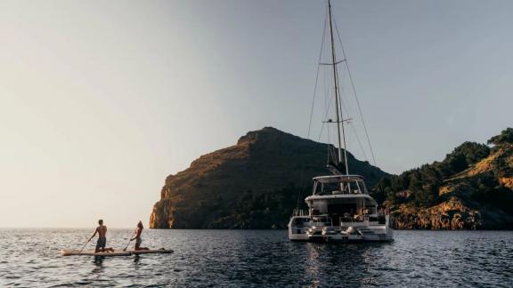 Two people paddle near the anchored catamaran in a bay with mountain views.