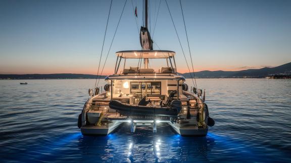 Among catamaran rental options in Kastela Triwing Ossus aft deck is seen with lights on anchored in calm sea at evening.
