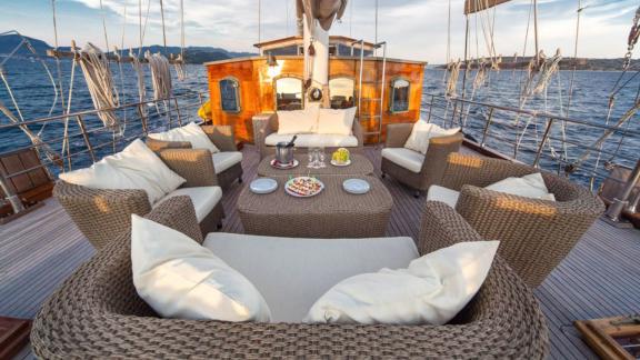 A gulet with a cosy seating area on deck, surrounded by calm sea and mountains in the background.