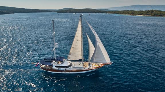 Gulet Lotus sails under full sail through the clear blue waters of the Croatian Adriatic, with a distant coastline in th