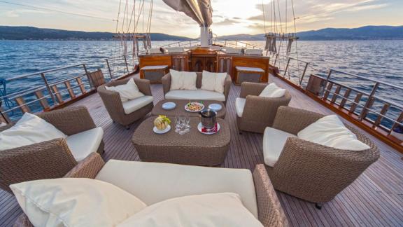 A gulet with a cosy rattan seating area on deck, set for a meal, surrounded by the calm sea and mountains in the backgro