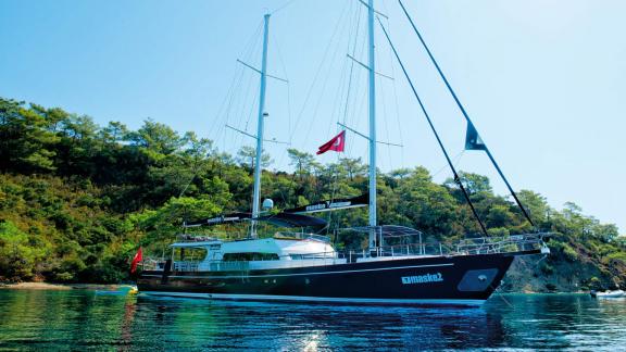 The charter yacht Maske 2 anchored before a green, wooded coastal landscape.