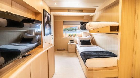 The cabin features upper and lower beds, a wide window, TV unit and light wood interior design.