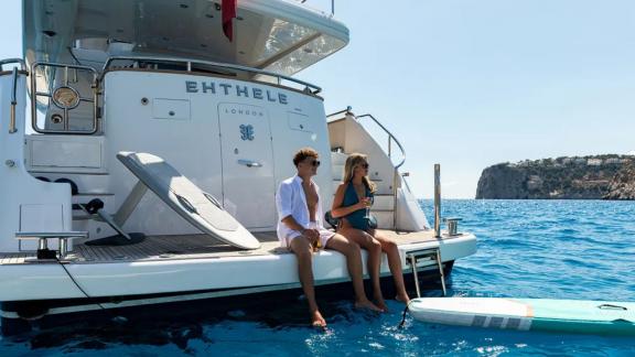 Two guests relax on the swim platform of Ehthele yacht anchored off Mallorca.