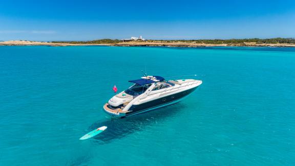 Anchored off Ibiza, the yacht has a paddleboard floating behind in clear blue sea.
