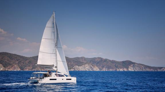Der Katamaran Pata segelt auf dem Meer, im Hintergrund ist eine Berglandschaft zu sehen.