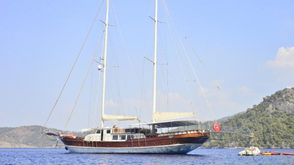 Gulet Berrak Su, arka planda berrak bir gökyüzü ile ormanlık bir sahilin açıklarında sakin sulara demirli.