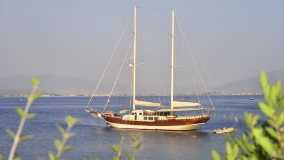 Sakin sularda demirlemiş iki direkli bir yelkenli, arka planda tepeler ve bir şehir.