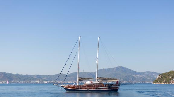 A gulet anchored on calm waters with mountains and a coastal town in the background.